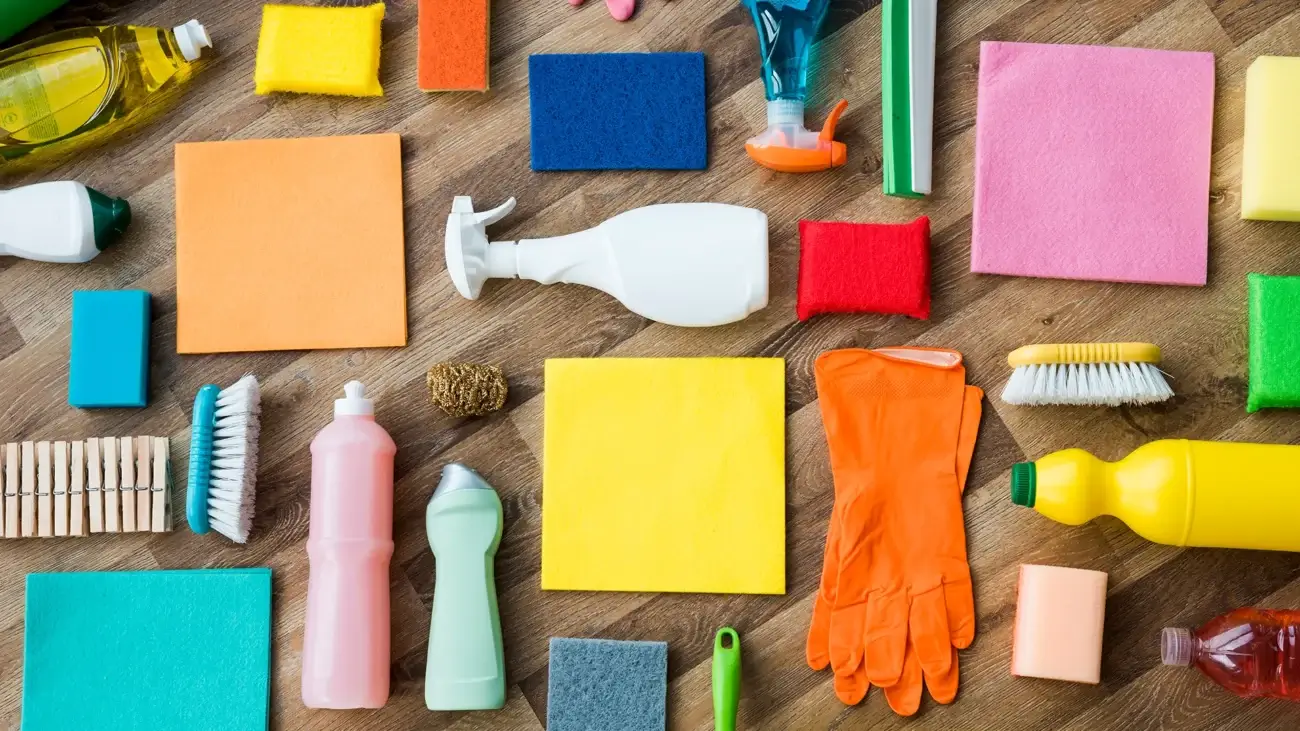 Assorted janitorial cleaning supplies and hygiene products neatly arranged on a wooden surface, including sponges, brushes, and gloves.